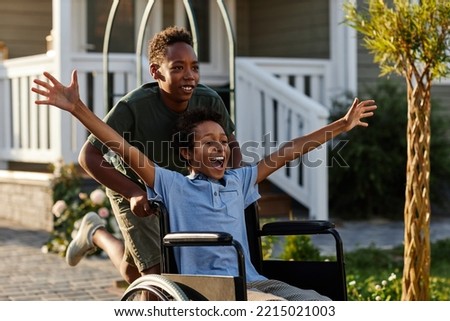 Portrait of black teenage boy pushing littlle brother in wheelchair while having fun together outdoors