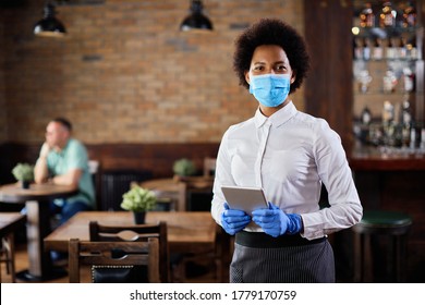 Portrait of black waitress wearing protective face mask while holding touchpad and looking at the camera in a pub. 