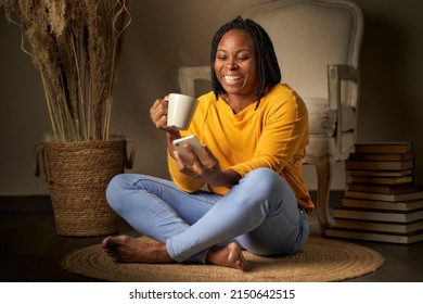 Portrait Of A Black Woman With Braided Hair Sitting On The Floor Drinking Hot Coffee While Looking At A Mobile Phone