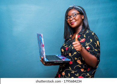 portrait of a black woman holding laptop and giving a thumbs up, isolated on a blue studio background-concept on technology and positive feedback