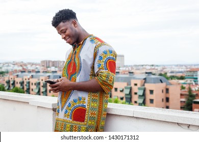 portrait of a black Young man wearing african traditional colorful clothes using smartphone while smiles on an european city rooftop background. Immigrant using phone to speak with far relatives.  