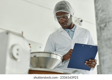 Portrait of black young man wearing lab coat and holding clipboard while overseeing production at food factory, copy space - Powered by Shutterstock