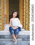 Portrait of A black-haired girl wearing a Thai dress with a silver sash sits smiling happily on the temple terrace while out sightseeing with her family.