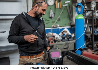 Portrait of a blacksmith during a break in a welding session. He takes the opportunity to smoke a cigarette, the welder's helmet can be seen in the bottom right. Blurred background with workshop. - Powered by Shutterstock