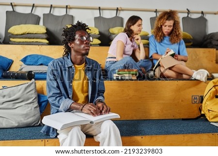 Portrait of blind African American man reading book in braille while sitting in library lounge, copy space