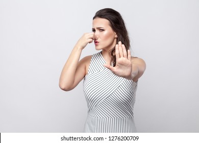 Portrait Of Blocking Woman Young Brunette Woman With Makeup And Striped Dress Standing With Stop Gesture And Holding Her Nose And Try To Stop Someone. Indoor Studio Shot, Isolated On Grey Background.