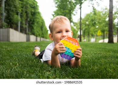 Portrait Blond Boy Lying On Grass And Playing With Toy Pop It. Stress And Anxiety Relief. Trendy Fidgeting Game. Developing Toy. Hand Motility. Outdoor Activities For Children. 