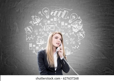 Portrait of a blond businesswoman talking on the phone while sitting near a blackboard with a round business sketch drawn on it.