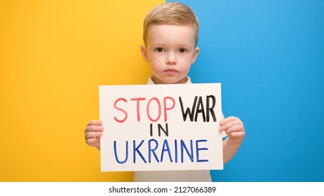 Portrait Blond little boy calls to Stop war in Ukraine, raises banner with inscription stop the war in Ukraine standing on blue-yellow studio background. No war, stop war, russian aggression.