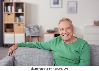 Portrait of a Blond Senior Man Sitting on a Gray Couch at the Living Area and Looking at Camera with Happy Facial Expression.