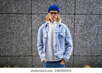 Portrait blond slim guy with long hair, scandinavian androgynous appearance in glasses looking at camera standing on street with hands in pockets. LGBT man gay transgender in denim clothes blue hat. 