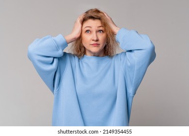 Portrait of blond stressed middle age woman hands on head, wear trendy blue sweatshirt, standing over light studio background