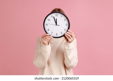 Portrait Of Blond Woman Holding Wall Clock Hiding Her Face, Time Management, Schedule And Meeting Appointment, Wearing White Sweater. Indoor Studio Shot Isolated On Pink Background.