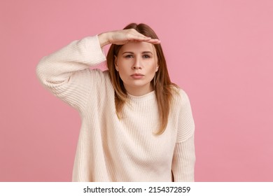 Portrait of blond woman peering into distance, looking far away with attentive view, searching on horizon and curious to discover, wearing white sweater. Indoor studio shot isolated on pink background