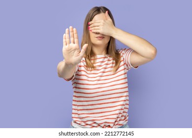 Portrait Of Blond Woman Wearing Striped T-shirt Showing Stop Gesture, Covering Eyes With Hand, Feeling Shamed And Scared To Watch. Indoor Studio Shot Isolated On Purple Background.