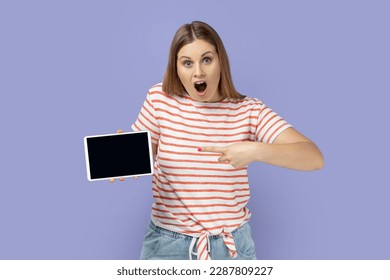 Portrait of blond woman wearing striped T-shirt pointing at tablet empty screen for adv, has unbelievable face, looking at camera with opened mouth. Indoor studio shot isolated on purple background.