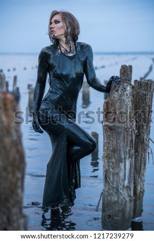 Portrait of Blonde Caucasian Girl Stays Smeared in a Healthy Black Mud in old Firth with Wooden Posts for Salt Production