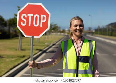 Portrait of a blonde Caucasian woman wearing a high visibility vest and holding a stop sign smiling to camera, standing in the road on a pedestrian crossing to help children cross the street safely - Powered by Shutterstock