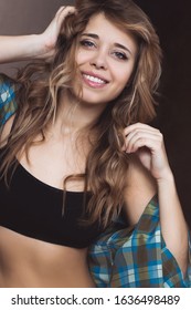 Portrait of a blonde caucasian young woman with a beautiful smile and lush wavy hair in a black short top and a checked blue shirt straightens her hair on a brown background. Real emotions