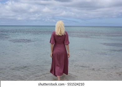 portrait of a blonde girl wearing a long purple dress standing in he ocean and touching the water