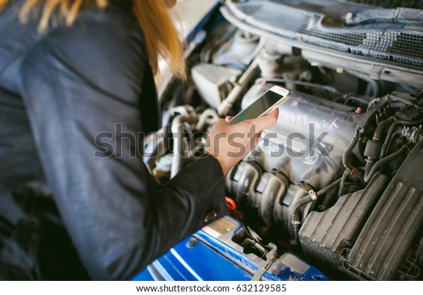 Portrait of blonde woman in black\
cloak calling phone in car service for help, lifting hood of car\
and looking at engine, malfunction, diagnosis,\
service