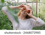 Portrait of blonde woman harvesting red ripe organic tomatoes in greenhouse and having fun. Healthy homegrown food concept. Cottagecore countryside