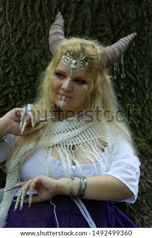 Portrait of a blonde woman with horns and pointed ears in the forest. White clothes. White geometric pattern on the face.