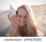 Portrait of blonde woman listening sea with seashell at beach.