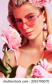 Portrait of blonde woman in pink sunglasses with pink peony around. Healthy skin and pink make up. Pink and purple background. Studio photoshoot                               