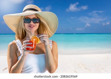 Portrait of a blonde woman with sunhat drinking a red cocktail drink in front of a tropical paradise beach and enjoying her summer holidays