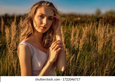 Portrait of a blonde young girl in a dress that sits in a field with tall grass against a background of blue sky and a tree. On a sunny day at sunset at golden hour in summer
