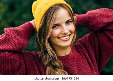 Portrait of a blonde young woman smiling broadly with a healthy toothy smile, wearing a red sweater, and yellow hat, posing on nature background in the park. People, travel, lifestyle