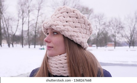 Portrait of a blue-eyed beautiful woman in white knitted hat and scarf standing in the winter city park and looking at the camera then turns her head in 4K slow motion close up video.