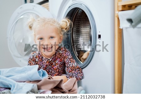 Portrait of blue-eyed girl with blonde hair tied in two buns cute sweet beautiful child is playing in pile of clothes colored for wash, smiles at camera in the background open washing machine