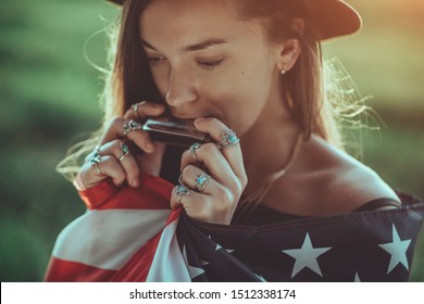 Portrait Of Boho Chic Woman In Hat With American Flag Wearing Silver Rings With Turquoise Stone Playes On Harmonica Outdoors. Jewelry Indie Girl With Hippie Style And Boho Fashion. Travel To America