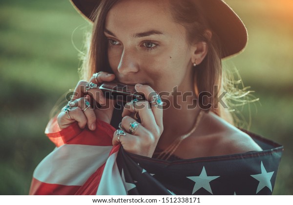 Portrait of boho chic woman in hat with american\
flag wearing silver rings with turquoise stone playes on harmonica\
outdoors. Jewelry indie girl with hippie style and boho fashion.\
Travel to america