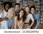 Portrait of bonding happy friendly young multiethnic college students embracing, posing in library near bookshelves. Group of joyful diverse millennial people looking at camera, relations concept.