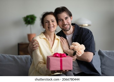 Portrait of bonding young grownup son and sincere joyful mature retired mother sitting on couch, holding bouquet of flowers and wrapped gift in hands, birthday or special occasion celebration.