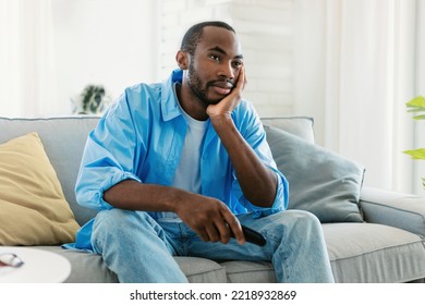 Portrait Of Bored African American Man Watching TV At Home, Sitting On Sofa And Leaning Head On Hand, Free Space. Discontented Black Guy Switching Channels With Remote Controller