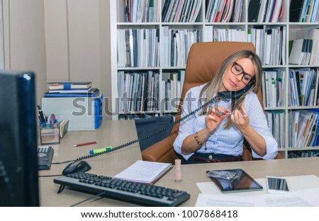 Portrait of bored blonde secretary polishing nails at workplace while talking on phone