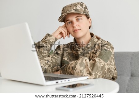 Portrait of bored Caucasian woman soldier wearing camouflage uniform and hat, sitting in front of notebook, keeping hand on her head, looking at camera with calm facial expression.