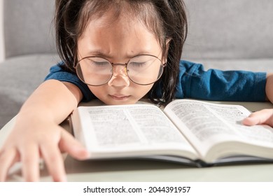 Portrait Of A Bored And Tired Little Toddler Asian Young Student Girl Wearing Glasses In A Blue T-shirt With A Textbook On The Desk At Home School. Education Learning And Child Future Success Concept.