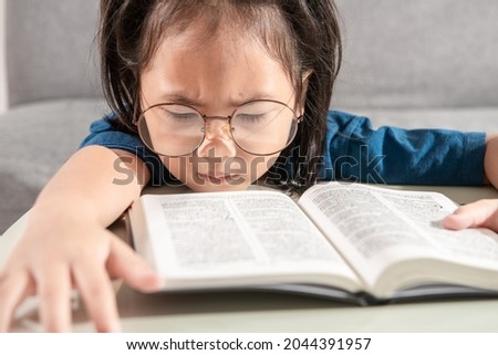 Portrait of a bored and tired little toddler Asian young student girl wearing glasses in a blue T-shirt with a textbook on the desk at home school. Education learning and child future success concept.