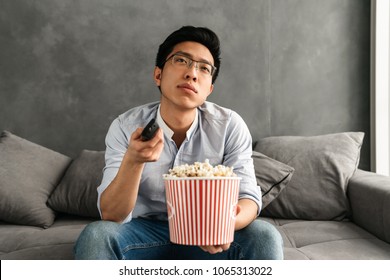 Portrait Of A Bored Young Asian Man Holding Popcorn And TV Remote Control While Sitting On A Couch At Home