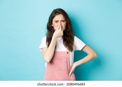 Portrait Of Bothered Cute Girl Shut Nose And Frown From Bad Smell, Smelling Terrible Reek, Something Stink, Standing Against Blue Background