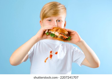 Portrait Of A Boy Biting Hamburger And Looking At The Camera. Isolated On Blue Background. Dirty Stain Tomato Sauce On White T-shirt. Daily Life Dirty Stain Concept. High Quality Photo