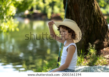 Portrait of a boy with curly hair in a straw hat sits by the pond