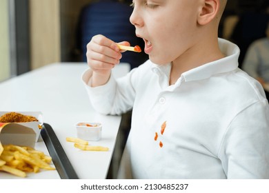 Portrait Boy Eating French Fries Potato Chips And Tomato Sauce At The Table In A Fast Food Restaurant. Dirty Stains Of Tomato Sauce On Clothes. Kids Eat Unhealthy Fat Food. 