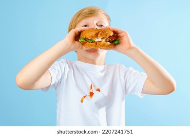 Portrait Of A Boy Eating Hamburger. Dirty Stain Tomato Sauce On White Clothes. Isolated On Blue Background. Daily Life Dirty Stain Concept. High Quality Photo