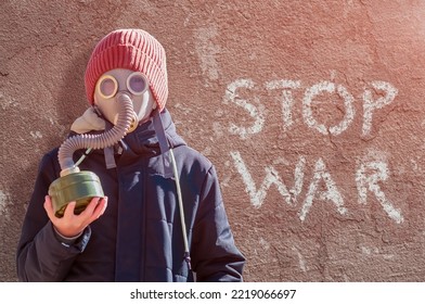 Portrait Of A Boy In A Gas Mask And Warm Clothes Against The Background Of A Red Old Plastered Wall. Bright Side Light. The Inscription On The Wall Stop The War. Concept - No War, World Peace.